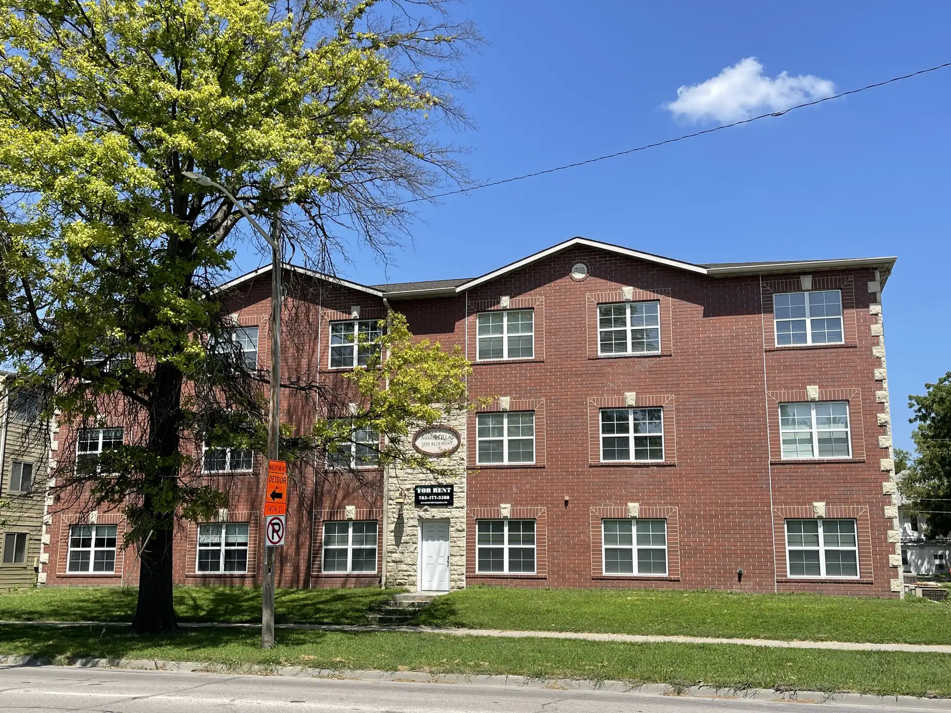 The four-story red brick building, known as Bluemont Villas, features white-trimmed windows and a small front entrance. Apartment 203 boasts views of the large nearby tree. A no-parking sign is visible on the street under the clear blue sky.