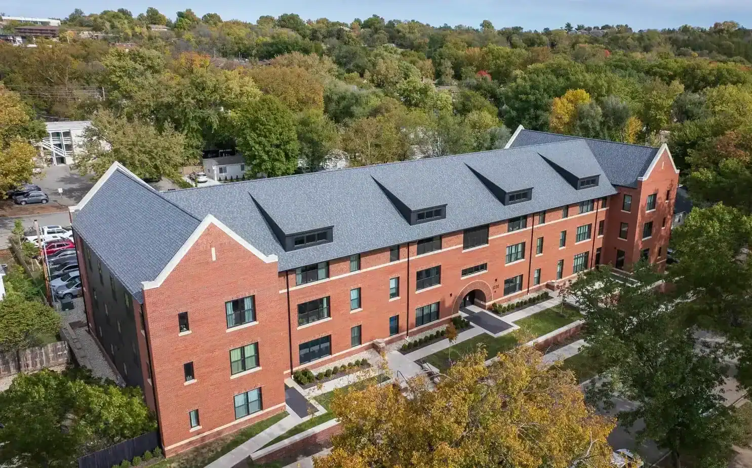 Aerial view of Apartment 1220, a three-story red brick building with a gray pitched roof, surrounded by trees in green and autumn colors, and a parking lot visible on the left&mdash;perfect for your next apartment rental.